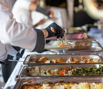 In a spacious and bright event hall, catering staff is actively serving a gourmet buffet. Guests are being offered various dishes, showcasing fresh vegetables and entrees.